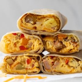 Stack of breakfast burritos cut in half to show fillings, including scrambled eggs, diced potatoes, red peppers, and sausage, with visible cheese. They are arranged on a white surface with scattered cheese shreds nearby.