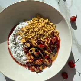 A breakfast bowl with cottage cheese, chia seeds, chopped cherries, peanut butter drizzle, and crunchy cereal