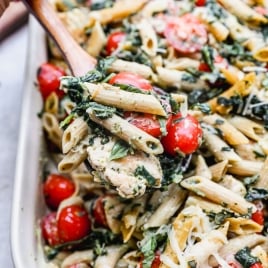 A wooden spoon scoops up baked chicken pesto pasta mixed with cherry tomatoes, shredded cheese, spinach, and herbs from a large white baking dish.