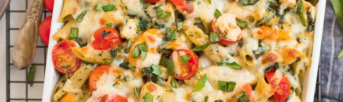 Baked pasta casserole with melted cheese, cherry tomatoes, and fresh basil in a white dish on a cooling rack.
