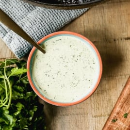 A small bowl of cilantro lime crema, a spoon inside, placed on a wooden surface near fresh cilantro, a gray towel, and a skillet with grilled chicken and lime.
