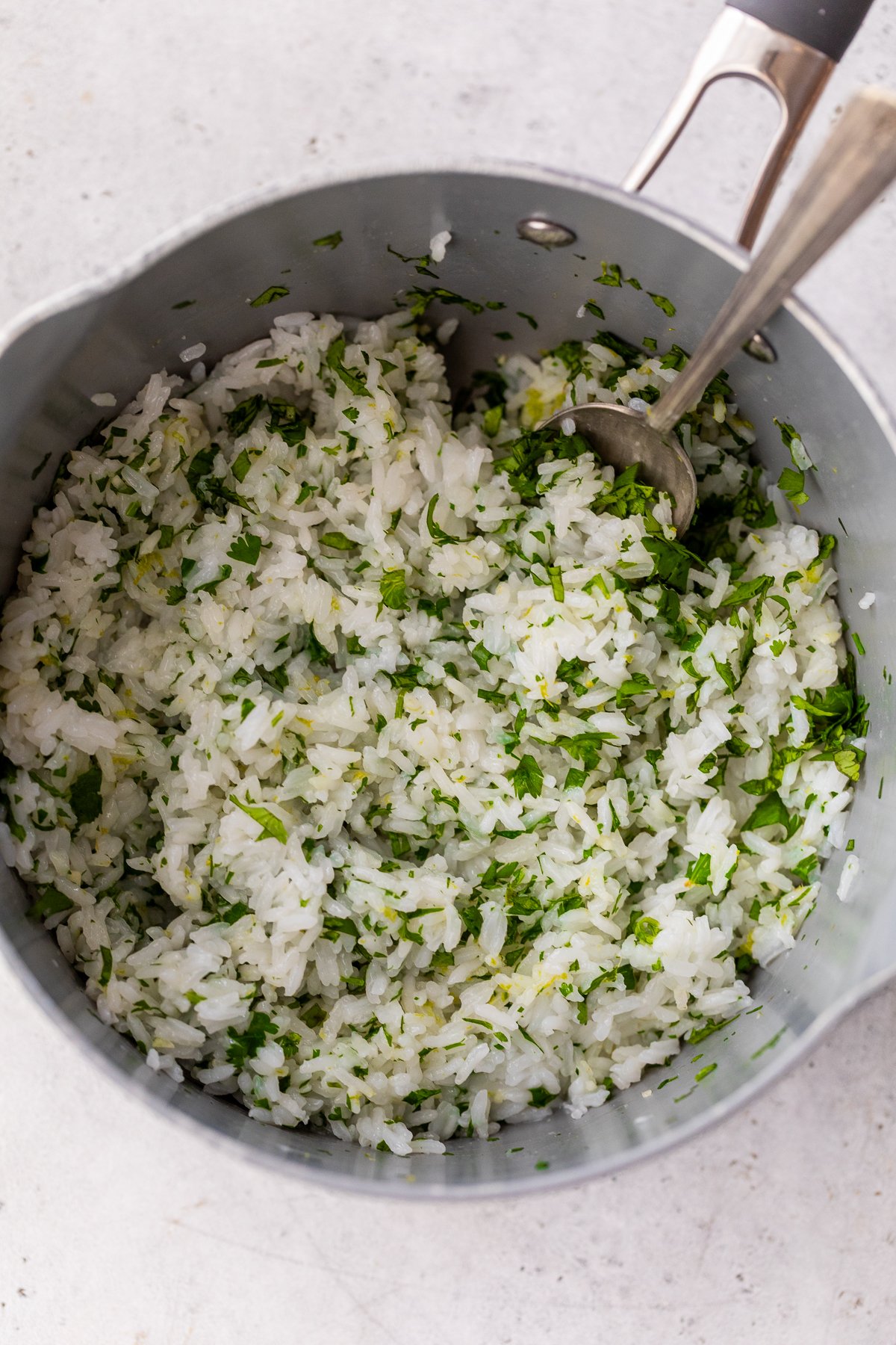 A pot filled with cooked cilantro lime rice, with a metal spoon resting inside, on a light-colored surface.