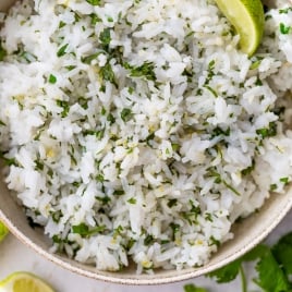 A bowl of cilantro lime rice, garnished with a wedge of lime. Extra lime wedges and fresh cilantro are on the table nearby.