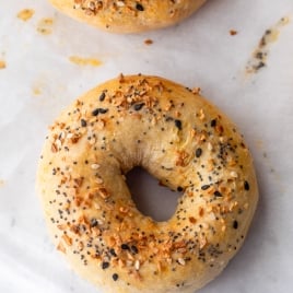 A close-up of a round, golden-brown cottage cheese bagel topped with a mixture of sesame seeds, poppy seeds, dried onion, and garlic on a sheet of parchment paper.