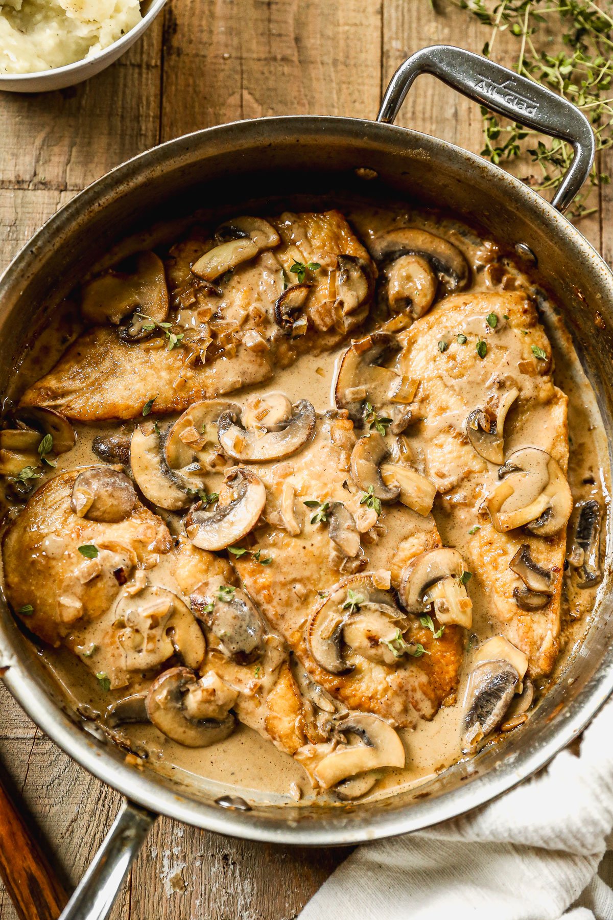 Mushroom chicken in a creamy sauce garnished with herbs, cooking in a metal skillet on a wooden surface.