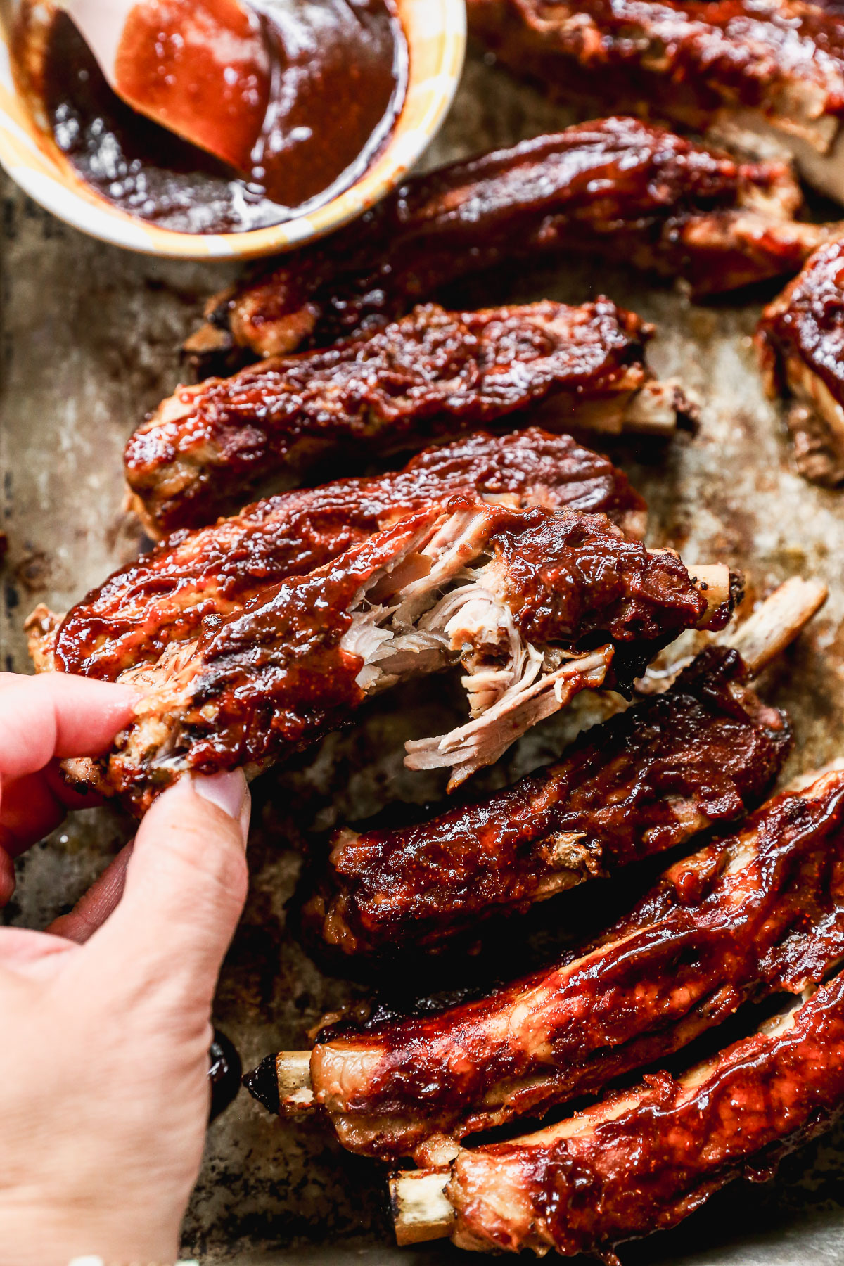 A hand pulls apart a tender, saucy barbecue crockpot rib from a tray filled with glazed ribs. A bowl of barbecue sauce and a brush are visible in the background.