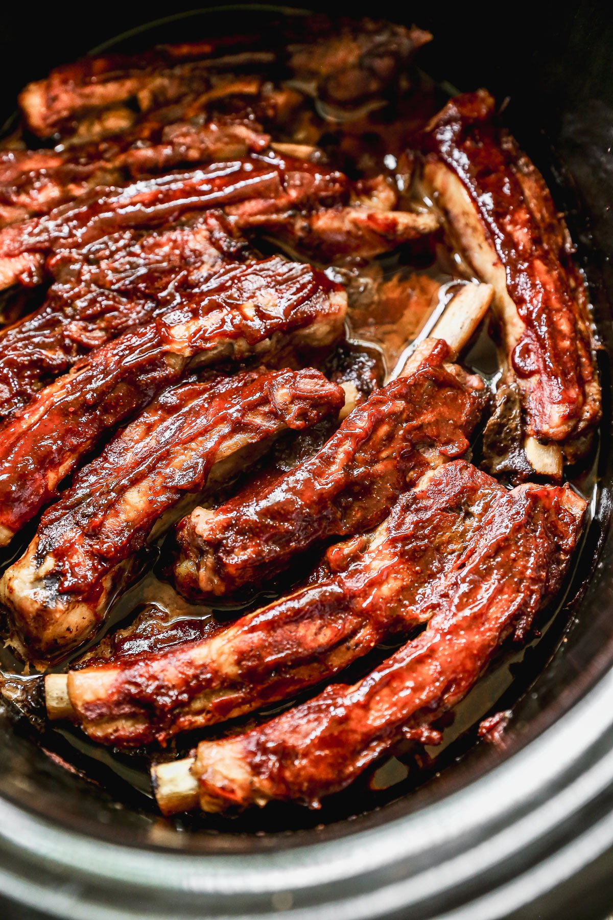 Barbecue pork ribs coated in sauce, slow-cooked in a crockpot, with meat glistening and slightly caramelized, arranged closely together in a dark, oval-shaped cooker.