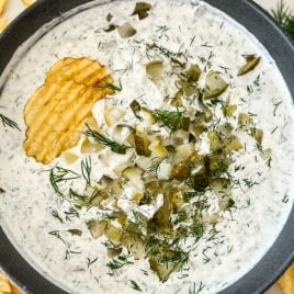 A bowl of creamy dip topped with chopped pickles and dill, surrounded by rippled potato chips.