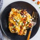 A slice of egg casserole with bacon, cheese, and herbs on a black plate with a fork, seen from above.