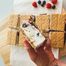 A hand holds a s'more ice cream sandwich with berries; more sandwiches and berries sit on a wooden board below.