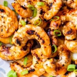 Close-up of grilled shrimp seasoned with herbs and spices, garnished with sliced green onions. The shrimp are slightly charred and arranged on a white plate.