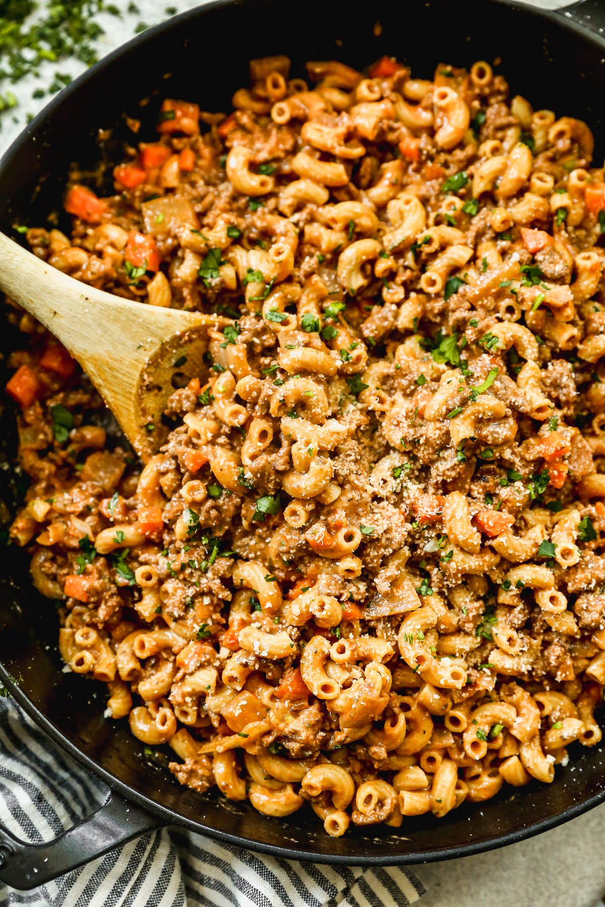 A skillet filled with ground beef pasta, topped with grated cheese. A wooden spoon is stirring the hearty pasta dish.