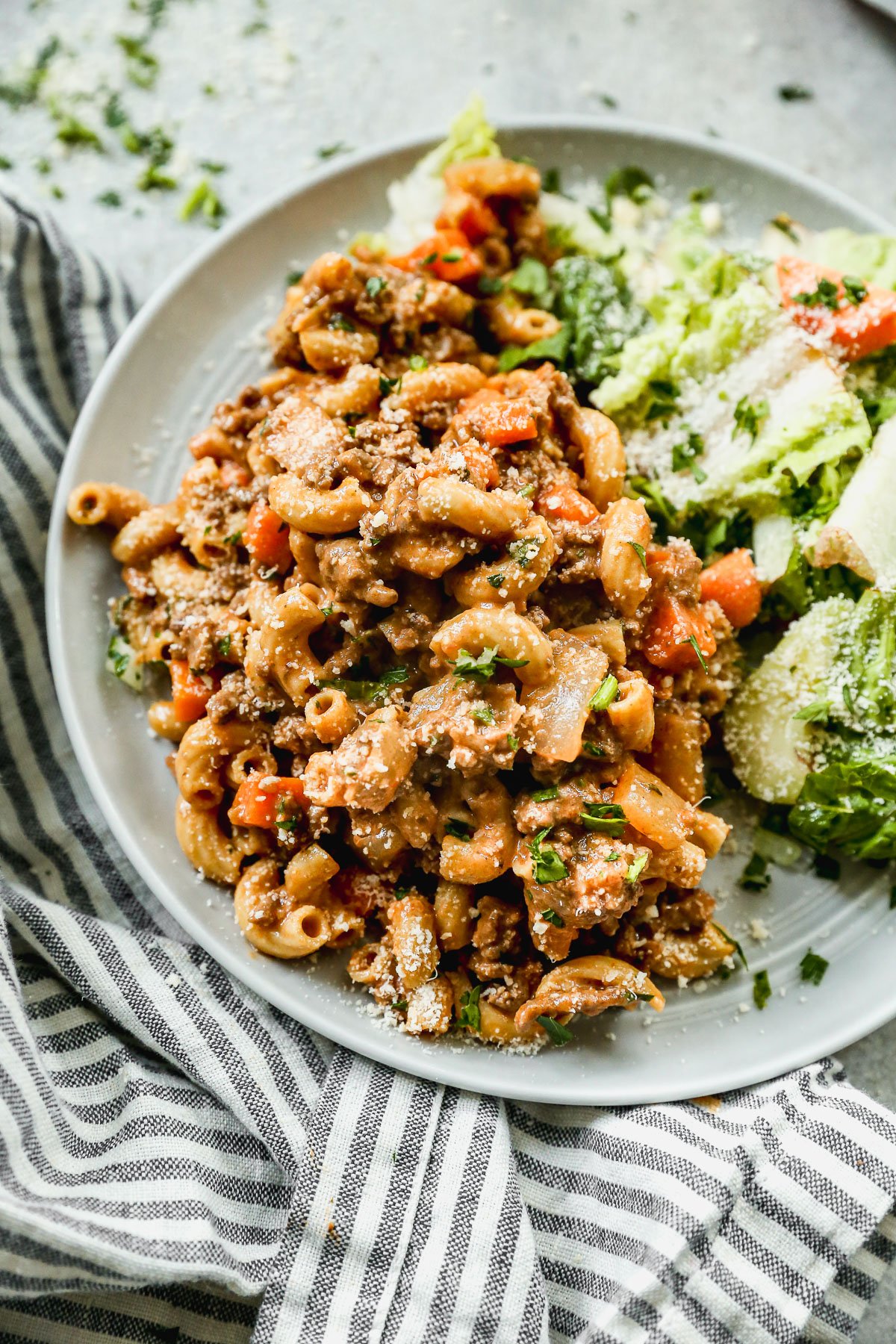 A plate of ground beef pasta with diced vegetables and grated cheese, served with a side of green salad and shredded parmesan, on a striped cloth napkin.