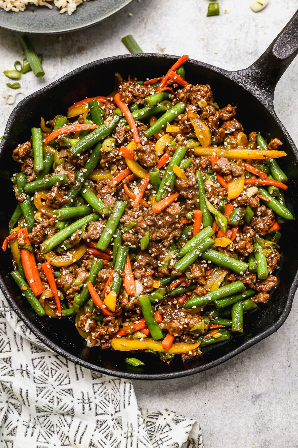 A skillet filled with stir-fried ground beef, green beans, red and yellow bell peppers, and carrots, garnished with sesame seeds and chopped green onions.