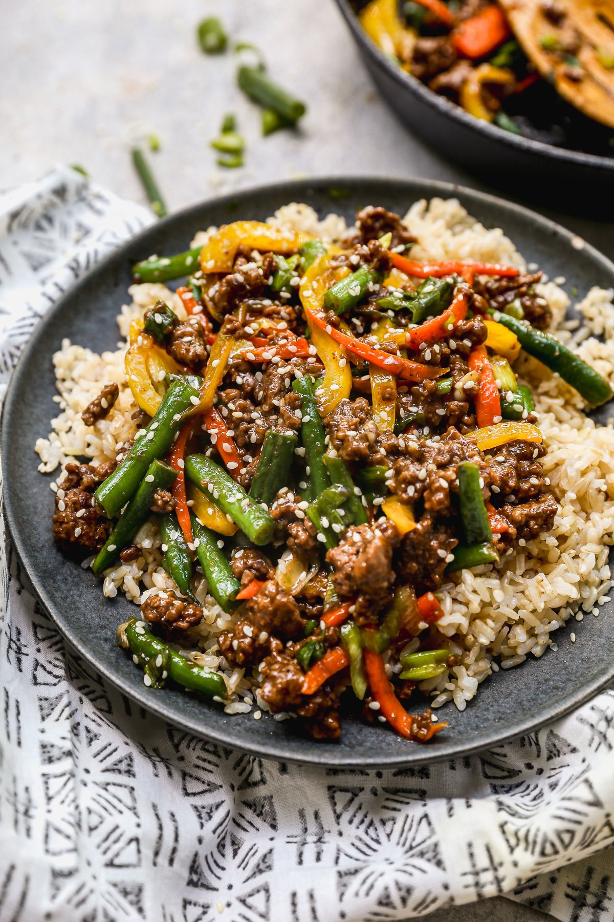 A plate of brown rice topped with ground beef stir fry with green beans, red and yellow bell peppers, all garnished with sesame seeds. The dish sits on a patterned cloth.