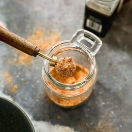 A wooden-handled scoop lifting spices from a glass jar. The jar is filled with hamburger seasoning mix. Other spice jars are blurred in the background on a grey countertop.