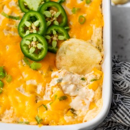 A baking dish filled with creamy, cheesy crab dip topped with melted cheddar, sliced green onions, and jalapeño slices. A potato chip is dipped into the dish, and a striped napkin is nearby.