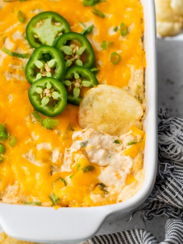A baking dish filled with creamy, cheesy crab dip topped with melted cheddar, sliced green onions, and jalapeño slices. A potato chip is dipped into the dish, and a striped napkin is nearby.