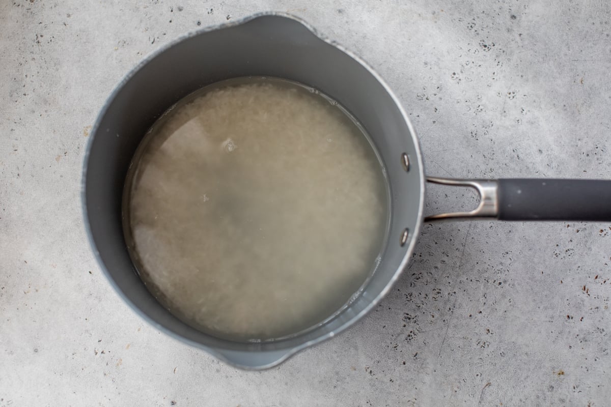 A grey saucepan filled with uncooked rice soaking in cloudy water sits on a light grey, textured surface.