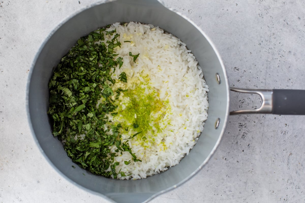 A silver saucepan filled with cooked white rice, chopped fresh cilantro, and grated lime zest on a gray surface.