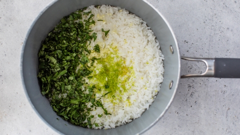 A silver saucepan filled with cooked white rice, chopped fresh cilantro, and grated lime zest on a gray surface.