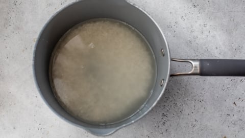 A grey saucepan filled with uncooked rice soaking in cloudy water sits on a light grey, textured surface.