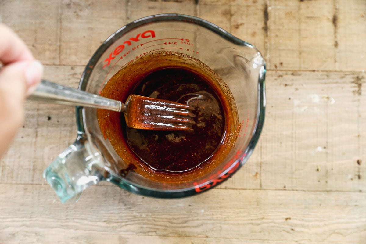 A hand uses a fork to mix a barbecue sauce in a glass Pyrex measuring cup on a wooden surface.