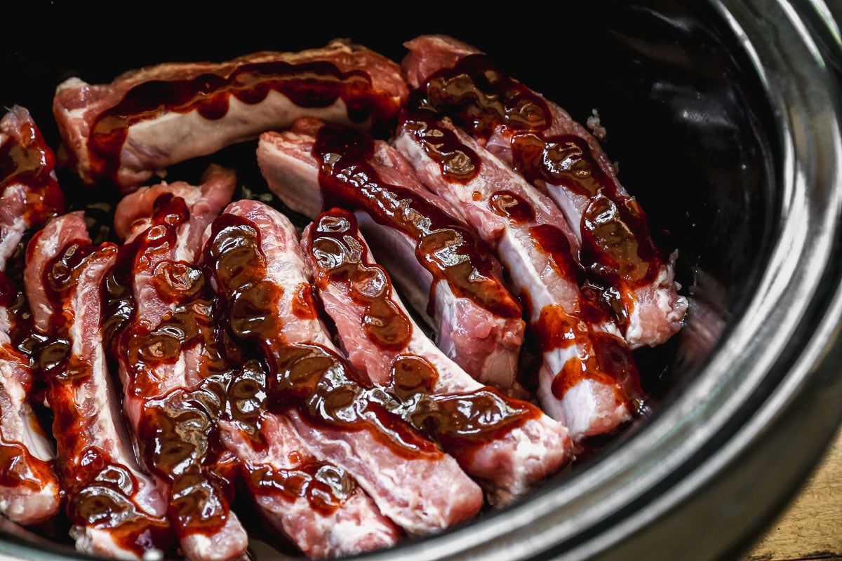 Raw pork ribs arranged in a slow cooker, topped with streaks of barbecue sauce ready for cooking.