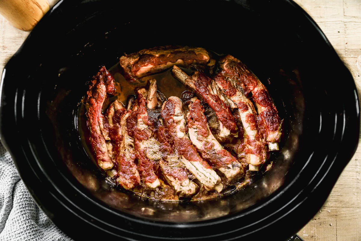 A slow cooker filled with cooked crockpot ribs arranged in a circle, sitting in their juices on the bottom; the ribs are browned and tender.