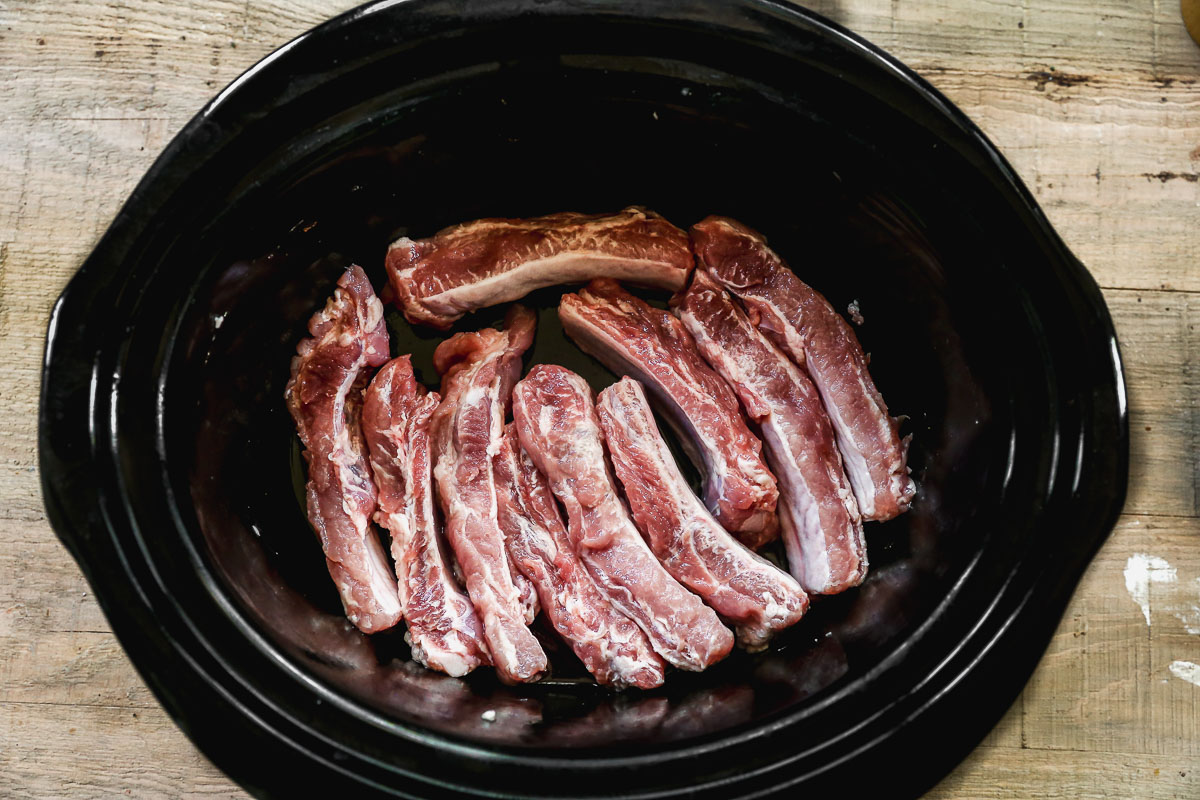 Raw pork ribs arranged in a single layer inside a black slow cooker, placed on a rustic wooden surface.