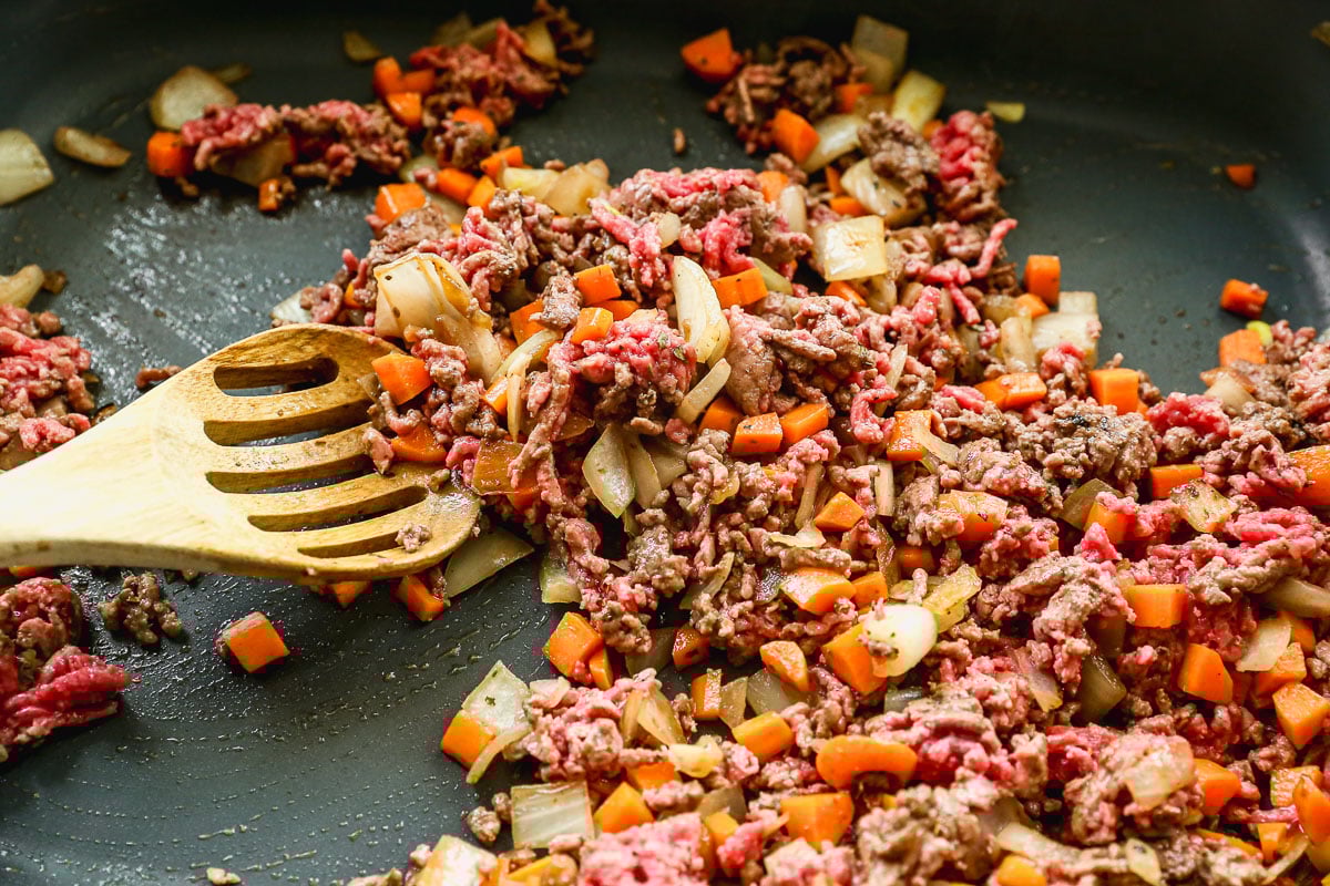 Chopped onions, carrots, and ground beef are being sautéed in a pan with a wooden slotted spoon, showing a mix of cooked and uncooked meat and vegetables.