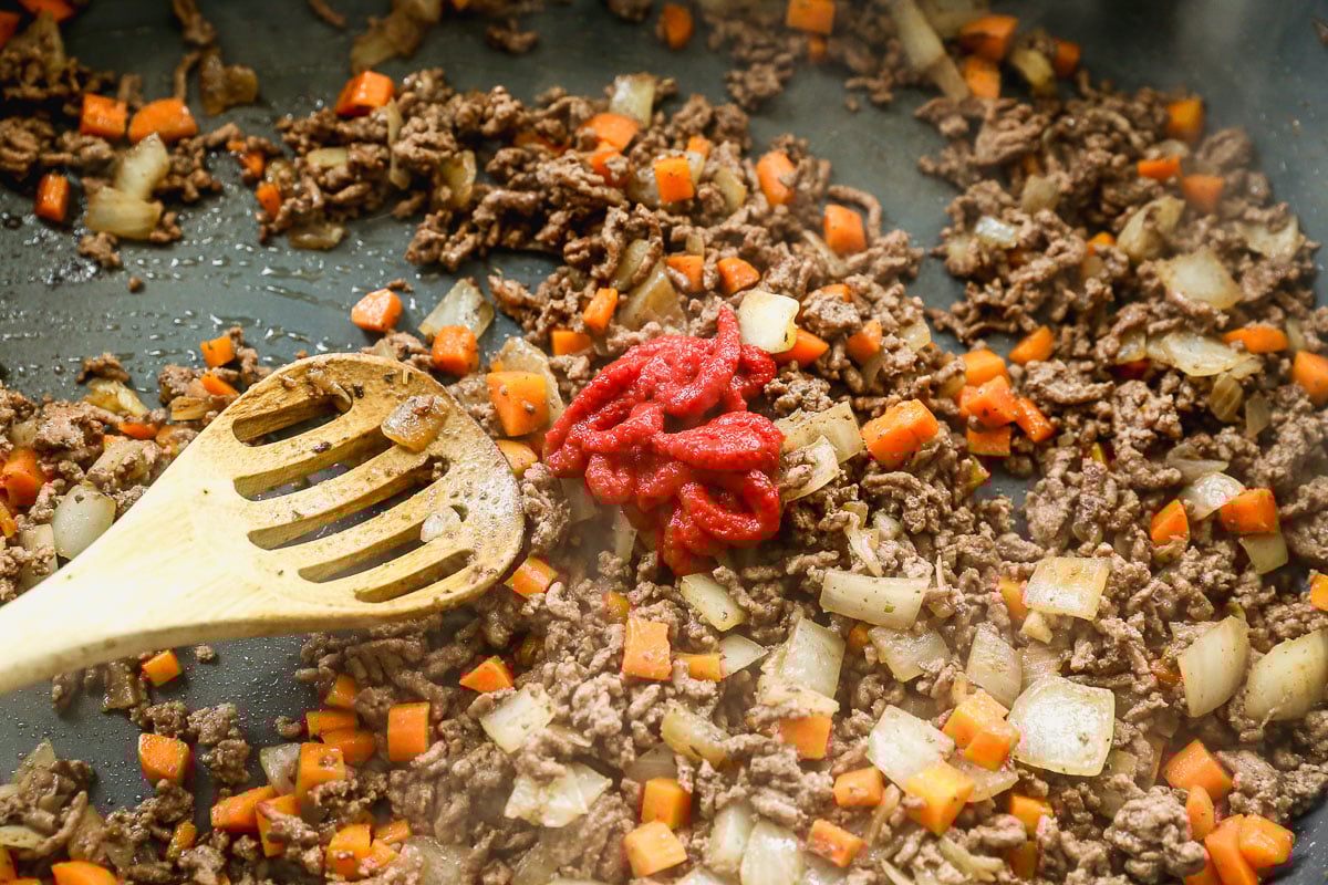 Ground beef, diced onions, and carrots are being cooked in a pan. A wooden spoon is mixing the ingredients, and a dollop of tomato paste sits on top of the mixture.