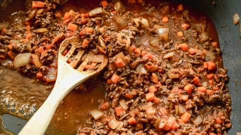 Ground beef, diced carrots, and onions simmering in a rich tomato sauce in a skillet, with a wooden spoon stirring the mixture.