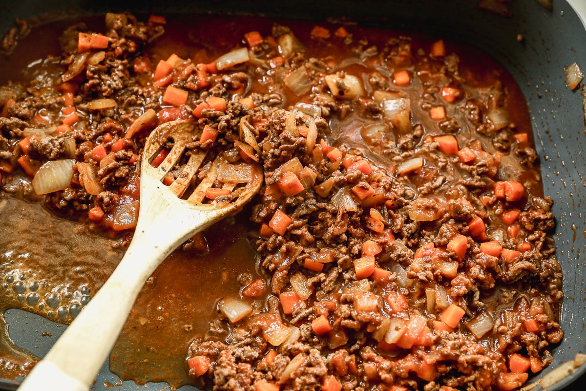 Ground beef, diced carrots, and onions simmering in a rich tomato sauce in a skillet, with a wooden spoon stirring the mixture.