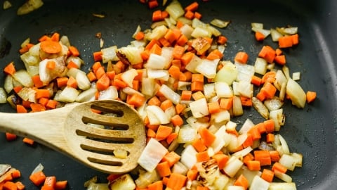 Chopped carrots and onions being sautéed in a pan with a wooden slotted spoon resting among the vegetables.
