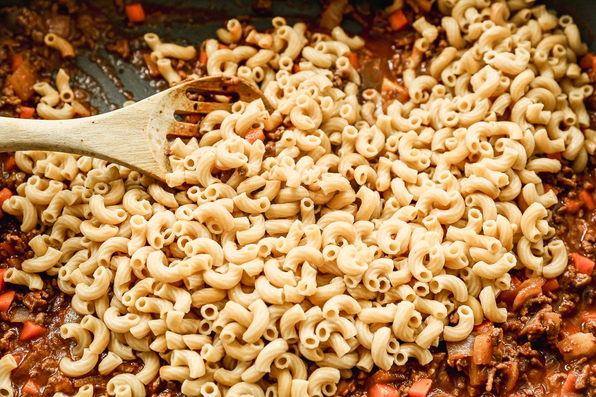 A wooden spoon rests on a skillet filled with cooked elbow macaroni, ground meat, red bell pepper pieces, and sauce, as the ingredients are being mixed together.