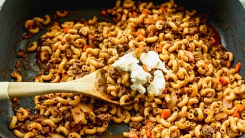 A skillet filled with cooked macaroni, ground beef, diced tomatoes, and onions, being stirred with a wooden spoon. There are dollops of cream cheese on top of the mixture.