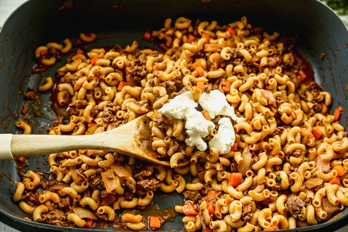 A skillet filled with cooked macaroni, ground beef, diced tomatoes, and onions, being stirred with a wooden spoon. There are dollops of cream cheese on top of the mixture.