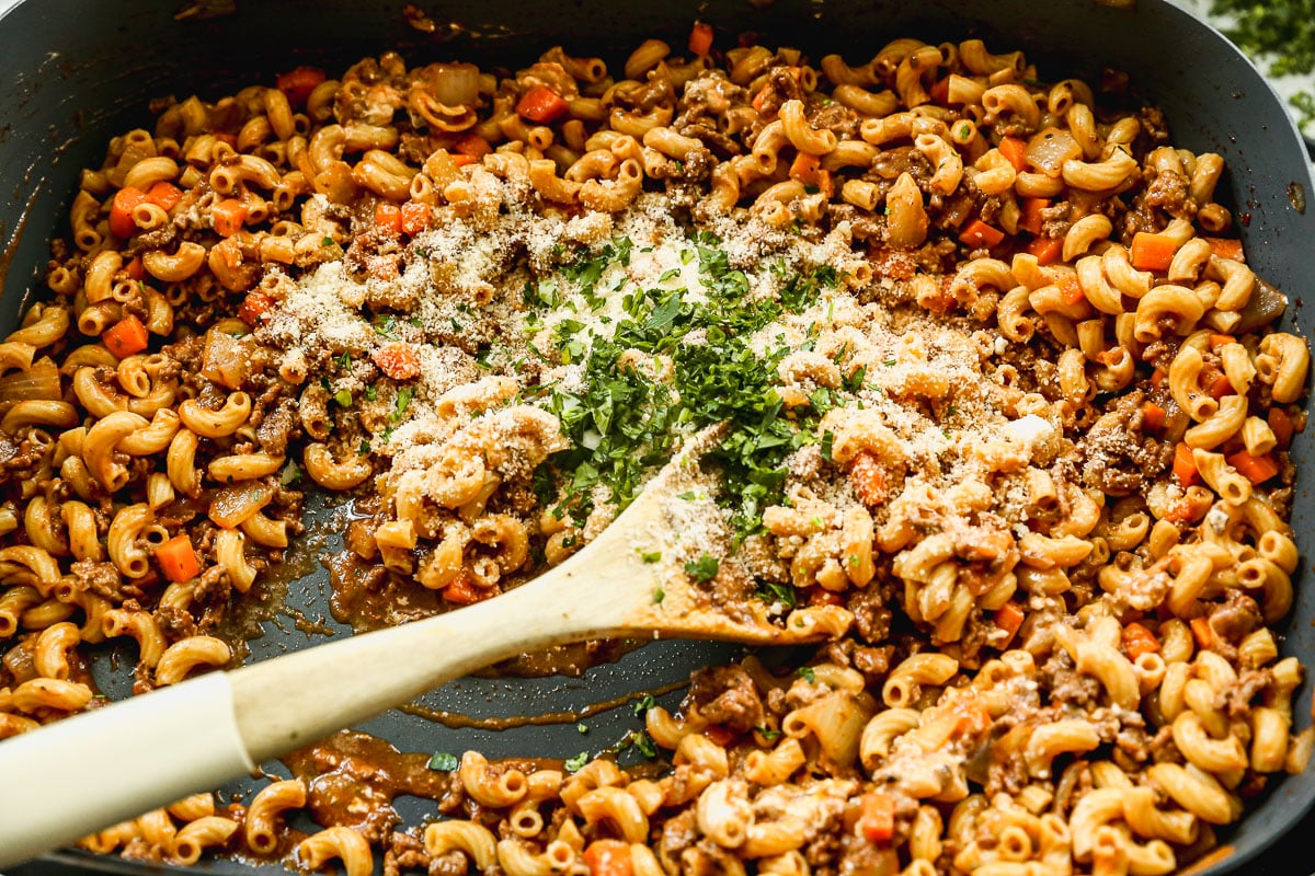 Elbow macaroni pasta with ground beef, carrots, tomato sauce, grated Parmesan cheese, and chopped parsley in a skillet. A wooden spoon rests in the mixture, ready for stirring.