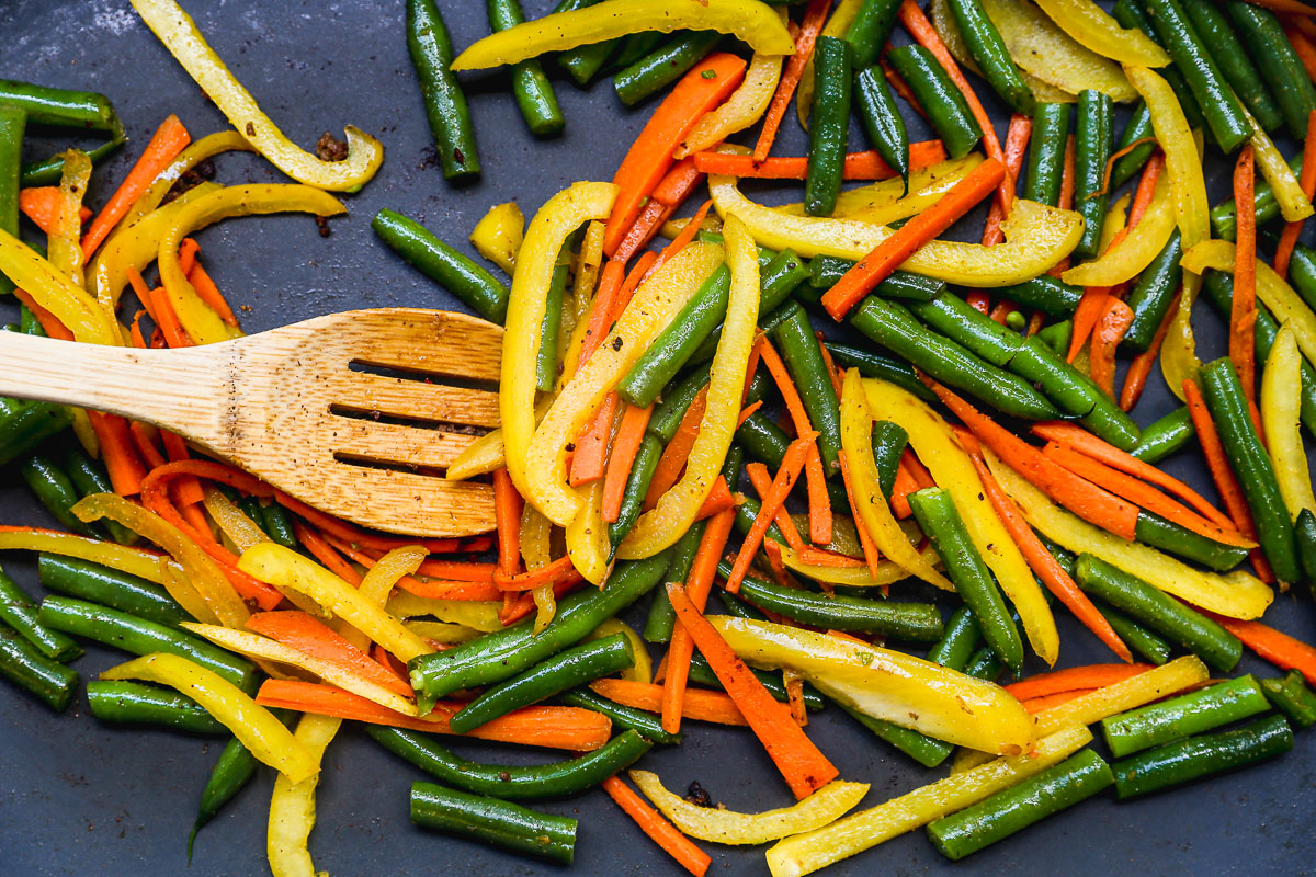Colorful sautéed vegetables for ground beef stir fry, including yellow bell peppers, green beans, and carrots, being stirred with a wooden spatula in a pan.