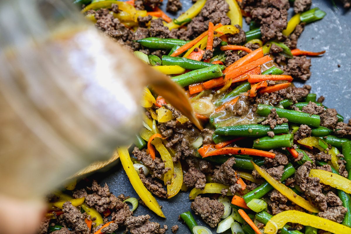 A close-up of stir fried ground beef, sliced green beans, yellow bell peppers, and carrots cooking in a pan, with sauce being poured in from a glass jar.