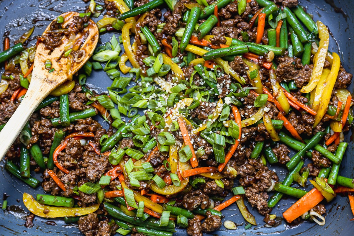 A close-up of a skillet with stir fried ground beef, sliced green beans, carrots, yellow bell peppers, and scallions, sprinkled with sesame seeds. A wooden spoon rests on the side of the pan.