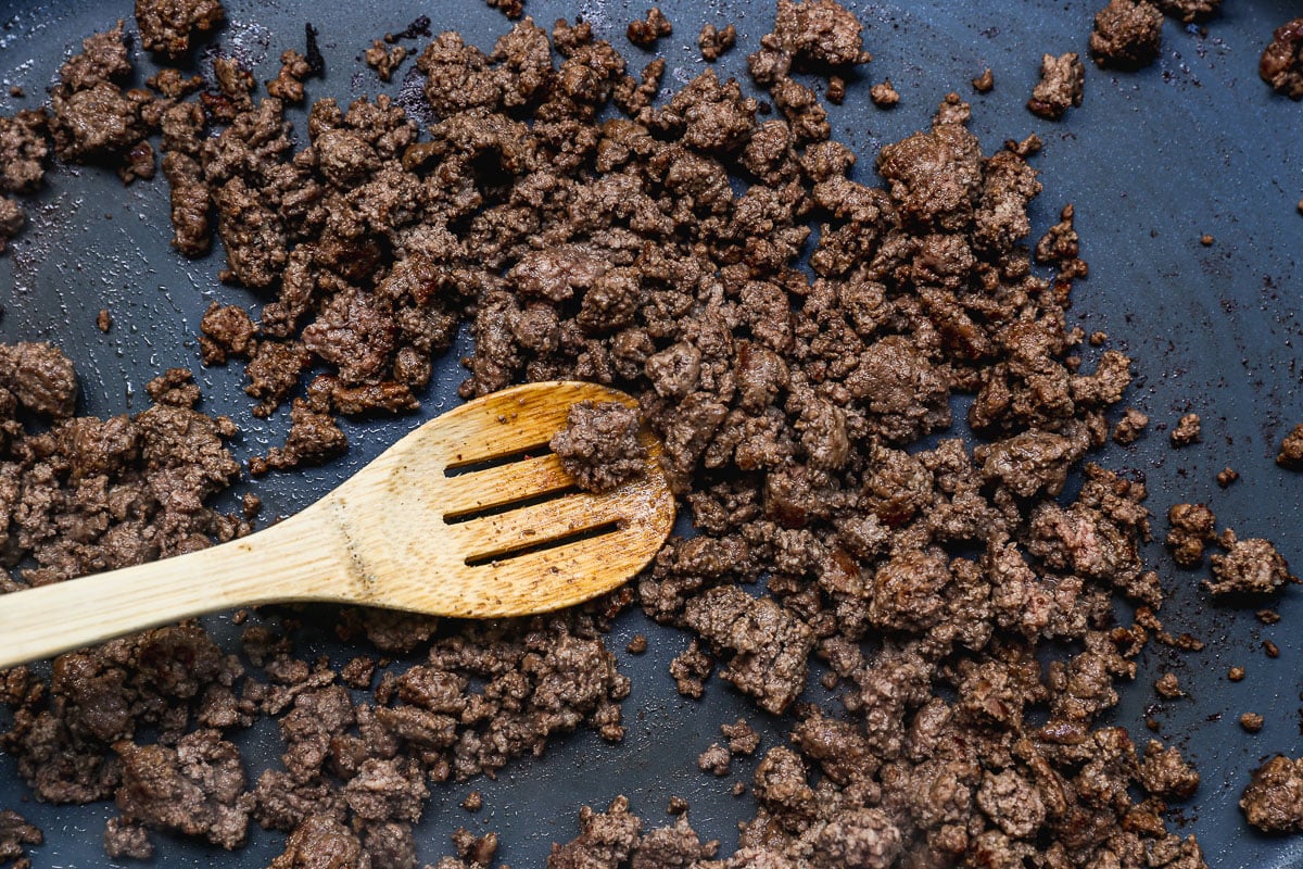 Cooked ground beef being stirred with a wooden slotted spoon in a dark nonstick skillet.