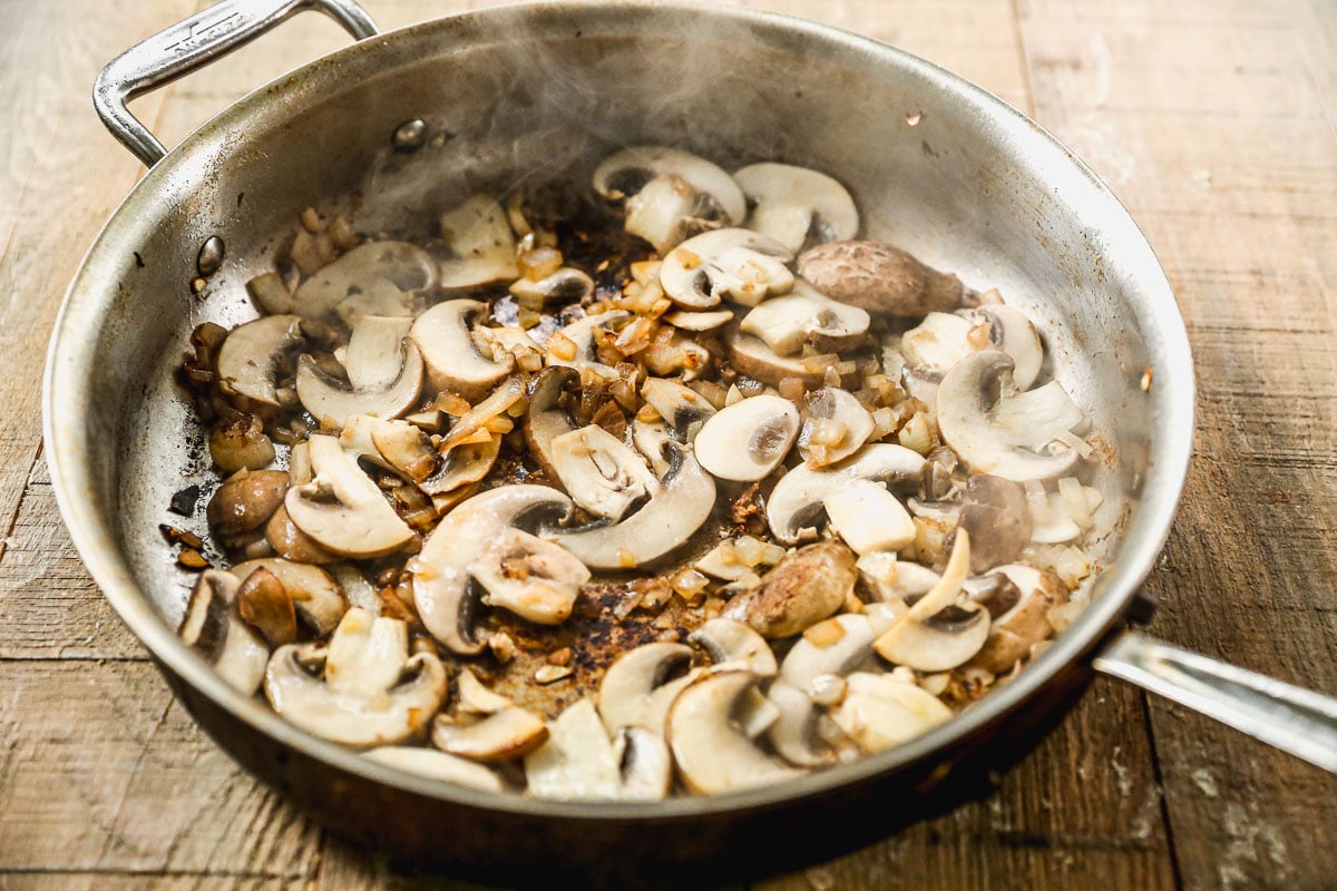 Sliced mushrooms and diced onions sautéing in a metal pan on a wooden surface, with visible steam rising.