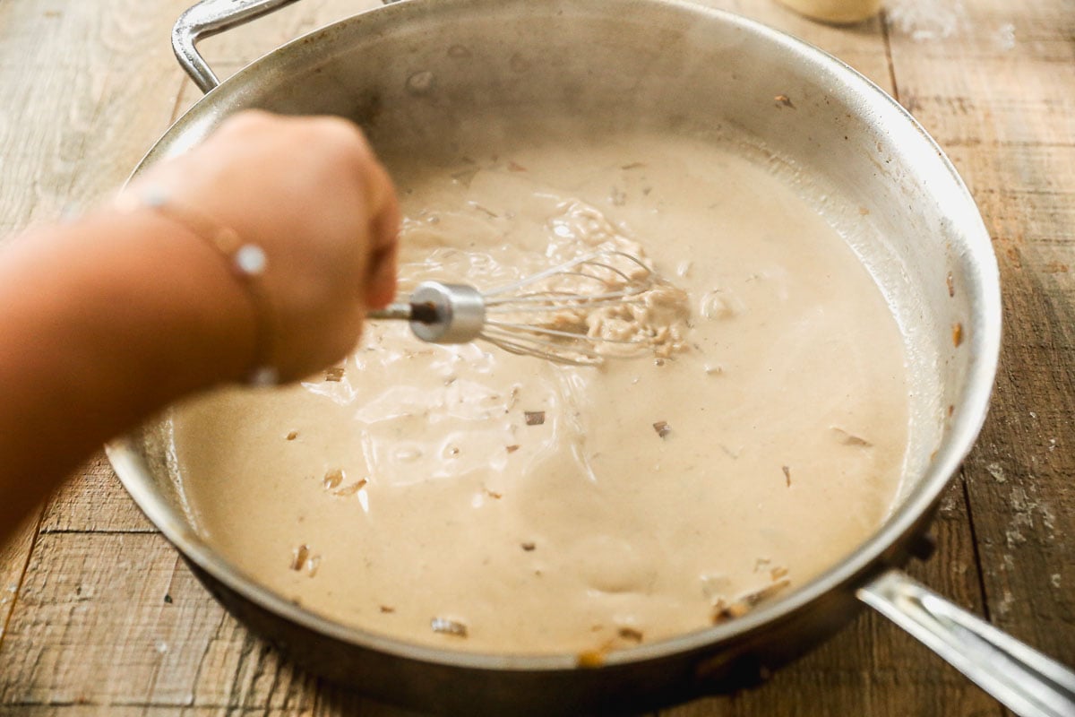 Hand whisking creamy sauce with onions in a large metal pan on a wooden table.