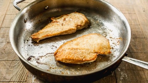 Two seared chicken breasts cooking in a metal skillet on a wooden surface.