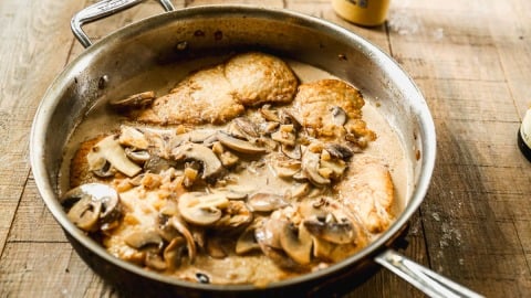 Chicken breasts and sliced mushrooms simmer in a creamy sauce in a metal skillet on a wooden surface.
