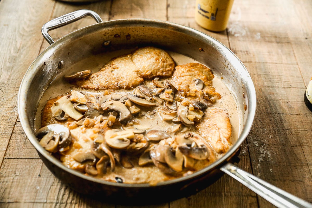 Chicken breasts and sliced mushrooms simmer in a creamy sauce in a metal skillet on a wooden surface.