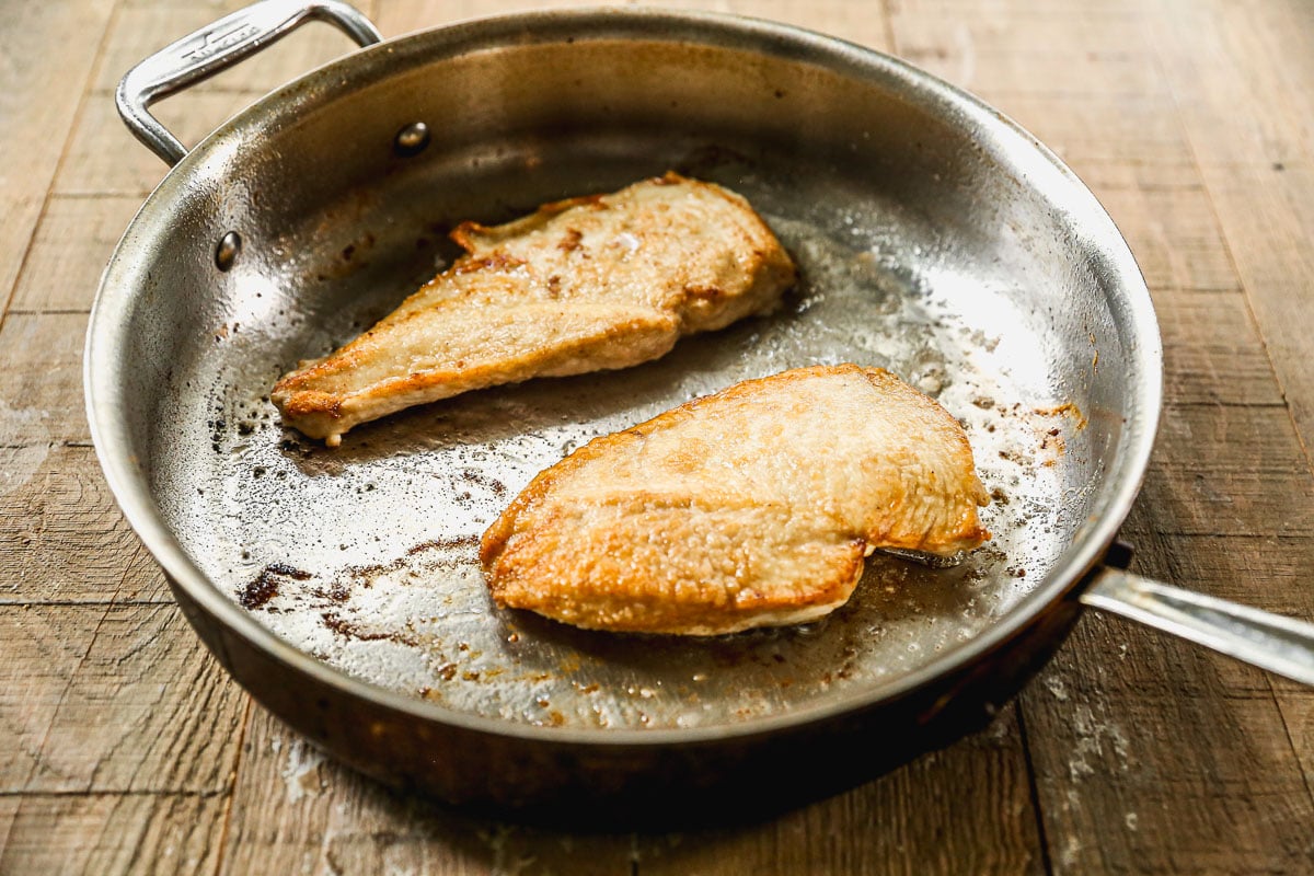 Two seared chicken breasts cooking in a metal skillet on a wooden surface.