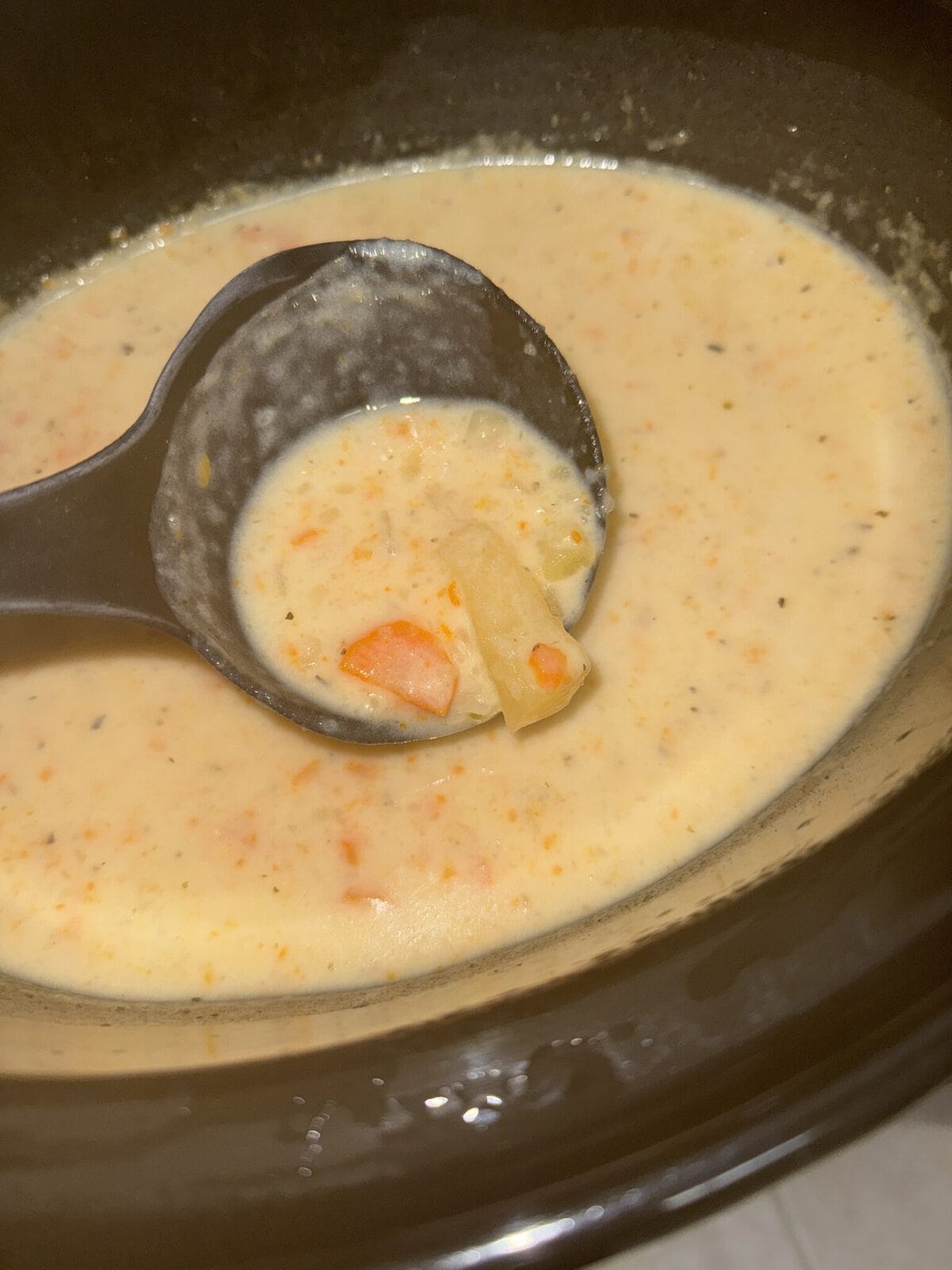 A ladle scooping creamy soup with visible chunks of carrots and potatoes from a pot.